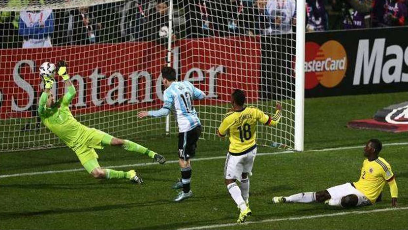 Jogo de futebol com dois jogadores da seleção da Colômbia, um deles ao chão, um jogador da Argentina acompanhando o movimento da bola em direção ao gol e salto do goleiro tocando a bola