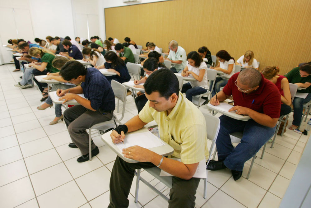 Pessoas em uma sala, sentadas em carteiras escolares, debruçadas sobre uma avaliação.