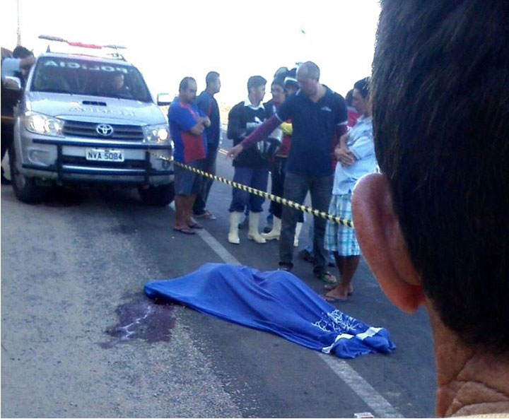 Corpo deitado no asfalto coberto com lençol, veículo da polícia e populares em volta.