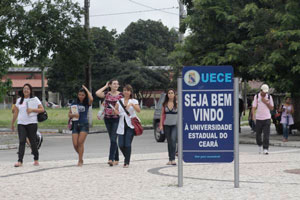 Jovens com livros no braço andando em calçada e em primeiro plano placa da UECE com letreiro de boas vindas.