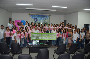 Várias pessoas posando para foto, onde muitas delas estão vestindo camisas da campanha outubro rosa e na frente segurando uma faixa com o nome da escola