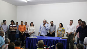 Prefeito Paulo Duarte, em pé, acompanhado de treza pessoas, falando ao microfone para um público sentado.