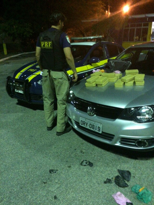 Carros da polícia federal e o utilizado pelo indivíduo perseguido, sendo o último com pacotes de pasta base de cocaína. Policial de costa entre os carros. 