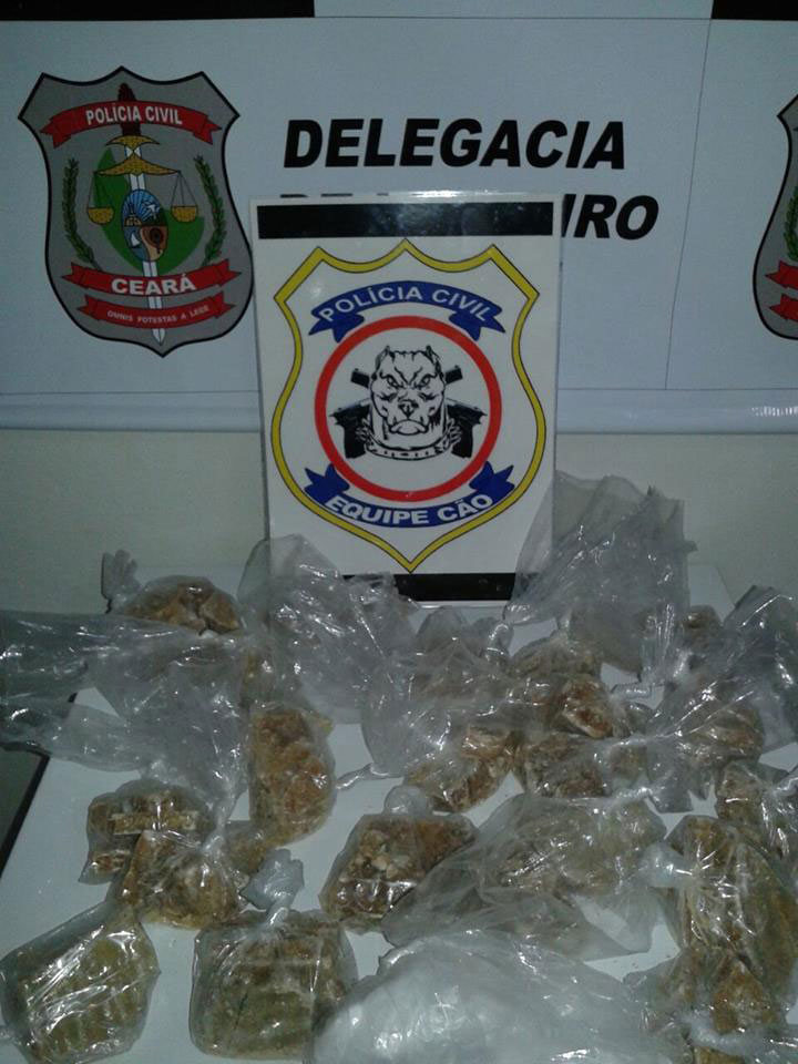 Pacote com drogras sobre mesa próxima a parede com os símbolos da Equipe ‘Cão’ da Polícia Civil de Limoeiro do Norte e Brasão da Polícia Civil do Ceará