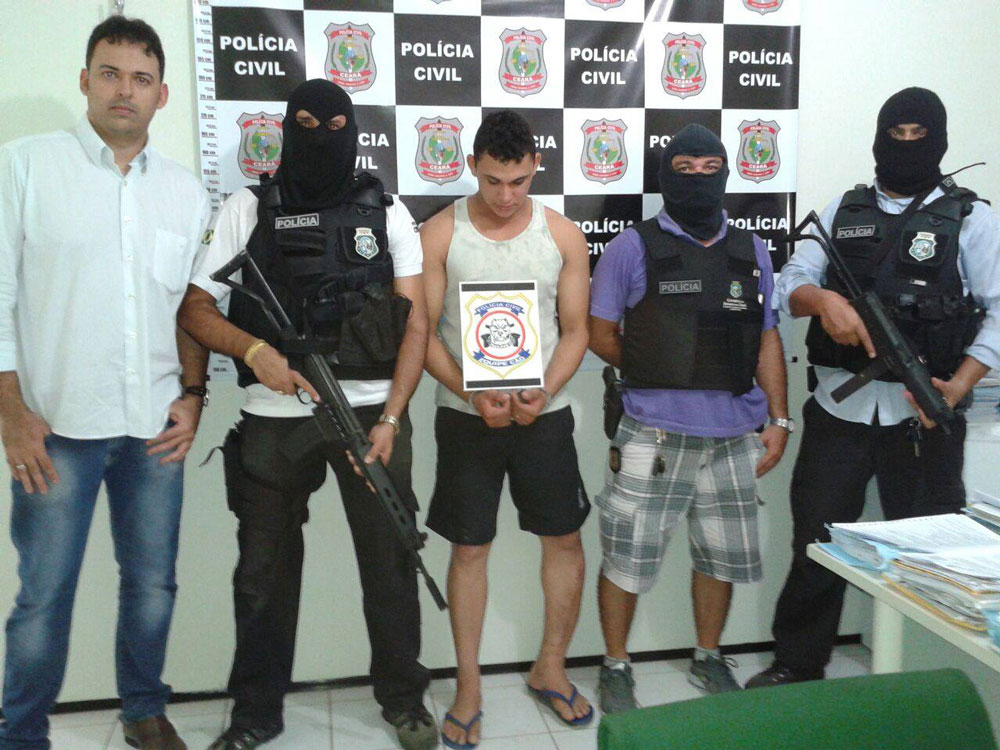 Foto composta por cinco pessoas em pé, sendo Francisco José de Assis Filho algemado no centro e agentes distribuídos igualmente ao seu lado armados e encapuzados com exceção de um.