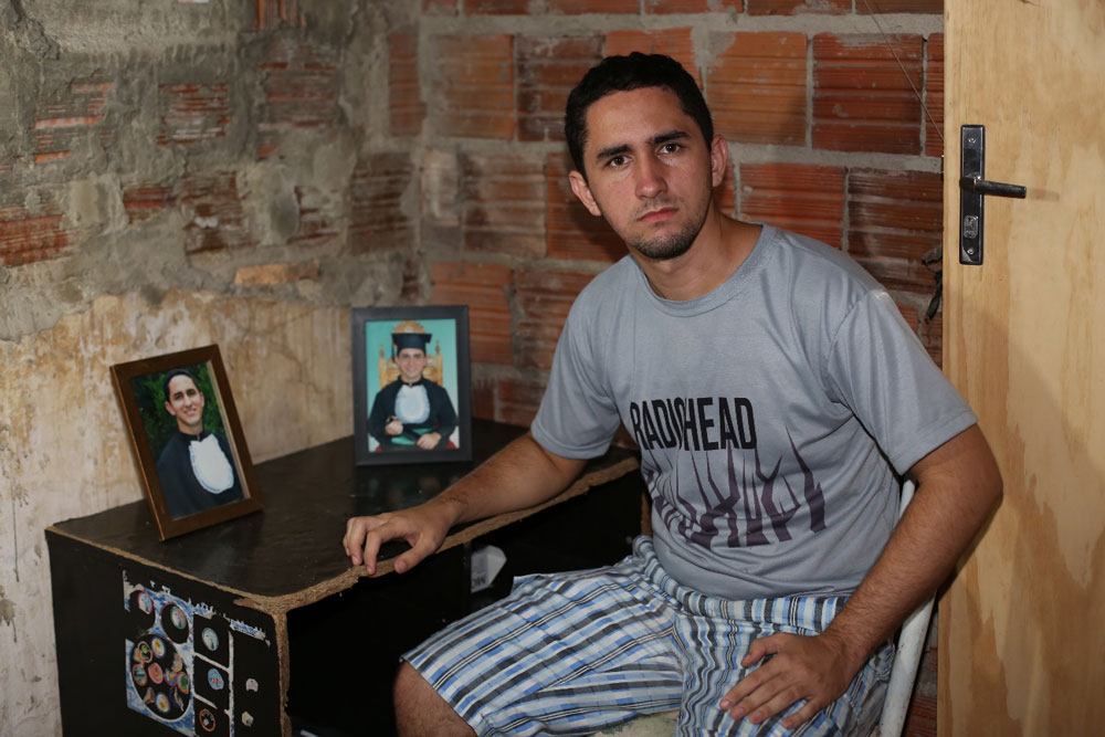 Adriel Pinheiro setando ao lado de uma mesa com dois porta retratos com suas fotos vestindo a beca de formatura.
