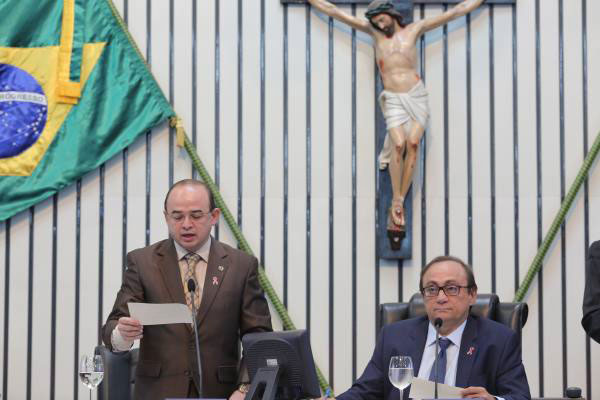 Dois homens trajando terno, um deles em pé enquanto lê um texto escrito em uma folha de papel. Ao fundo a bandeira do Brasil e uma imagem de Jesus crucificado.