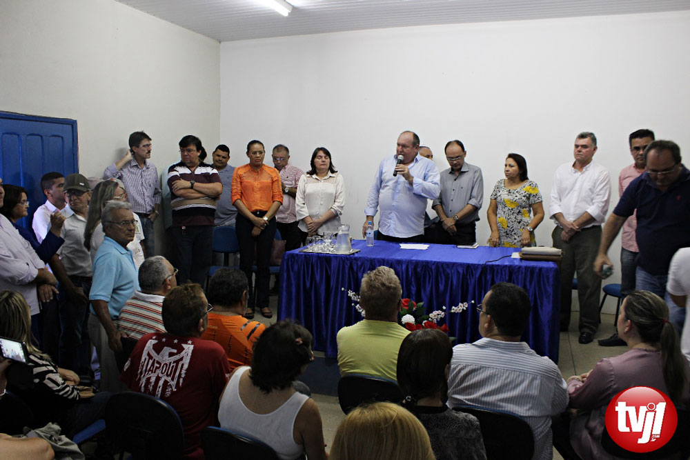 Prefeito Paulo Duarte, em pé, acompanhado de treza pessoas, falando ao microfone para um público sentado.