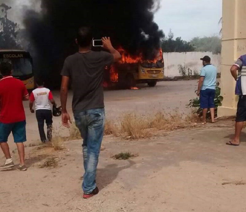 Pessoas em pé observando Ônibus em chamas ao fundo.