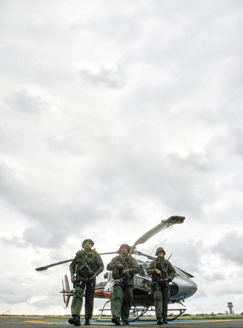Três soldados fardados e armados caminhando com helicóptero pousado ao fundo em dia nublado 