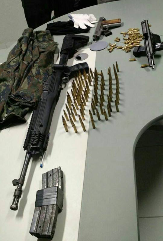 Um fuzil americano Rugger, calibre 5.56;  duas pistolas; uma farda camuflada e muita munição sobre uma mesa