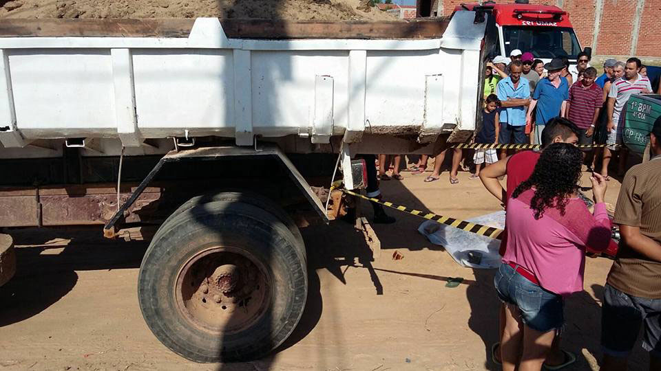 Visão parcial da traseira de um caminhão, fitas de contenção, pessoas em volta e corpo coberto ao chão