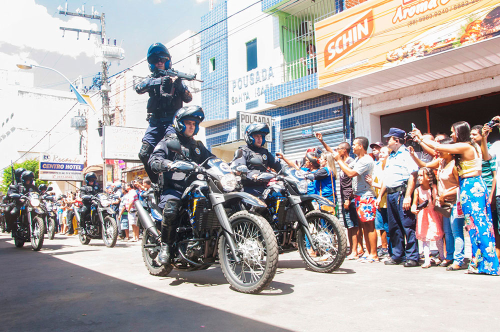 Policiais em motos percorrendo rua, com população a margem observando a passagem.