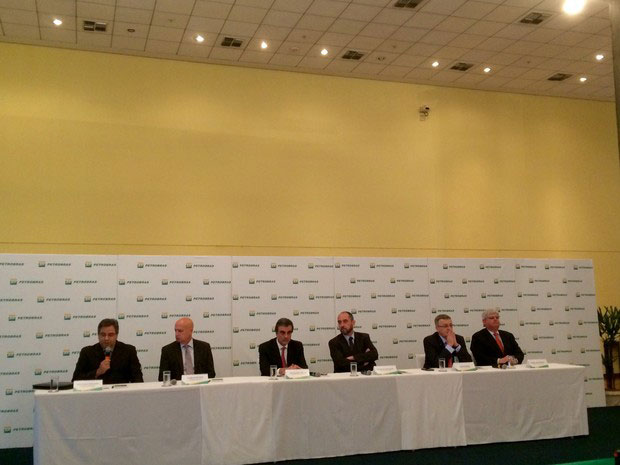 Mesa branca e larga composta por seis homens vestindo terno e ao fundo painel com estampado várias repetições do logotipo da Petrobras.