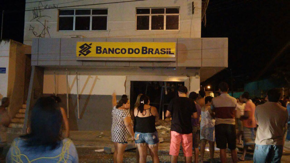 Fachada da agência do Banco do Brasil com destroços na porta de entrada e populares olhando.