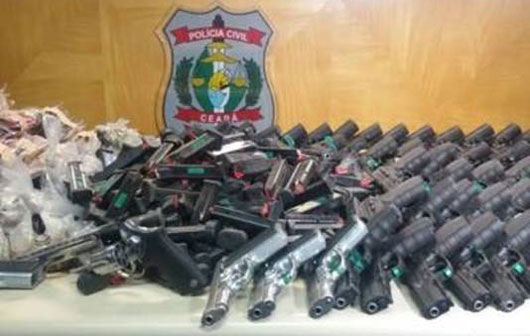Várias armas sobre uma pesa e ao fundo o brasão da Polícia Civil