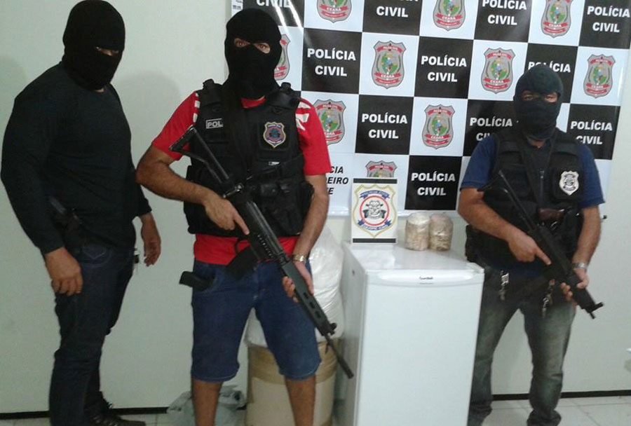 Três policiais com capuz preto e armados ao lado da droga apreendida