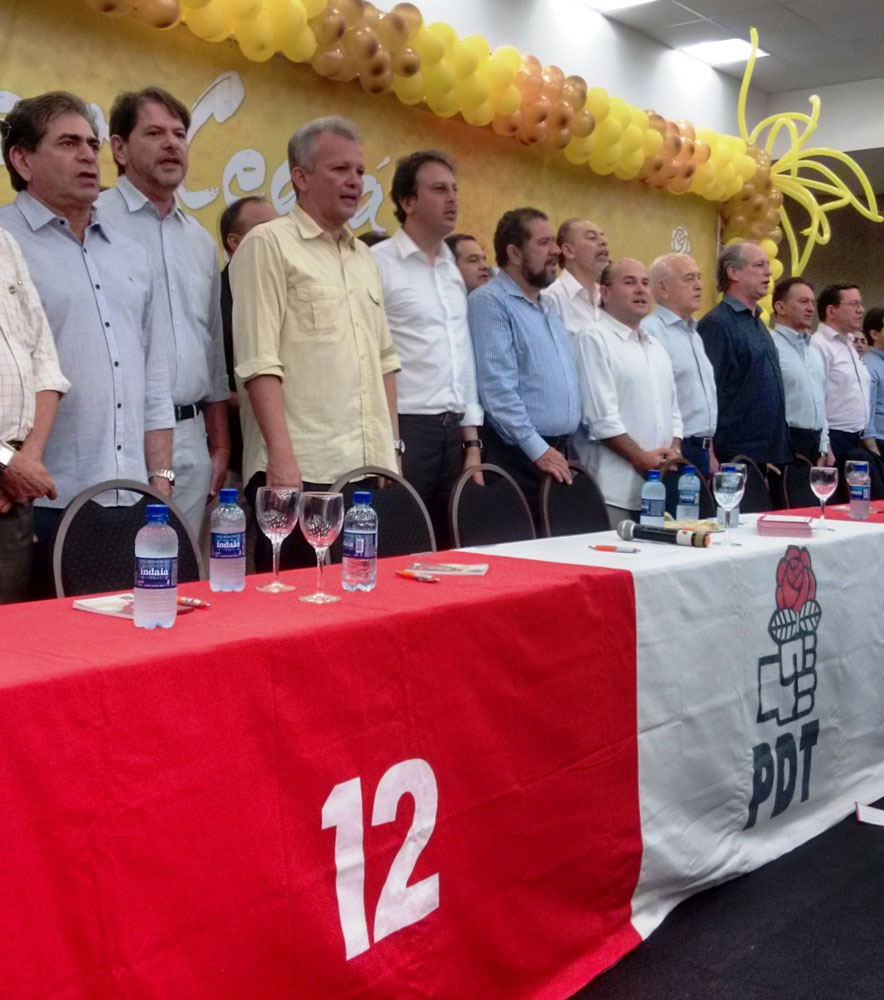 Vários políticos alinhados lado-a-lado atrás de uma longa mesa coberta com toalha personalizada do partido PDT