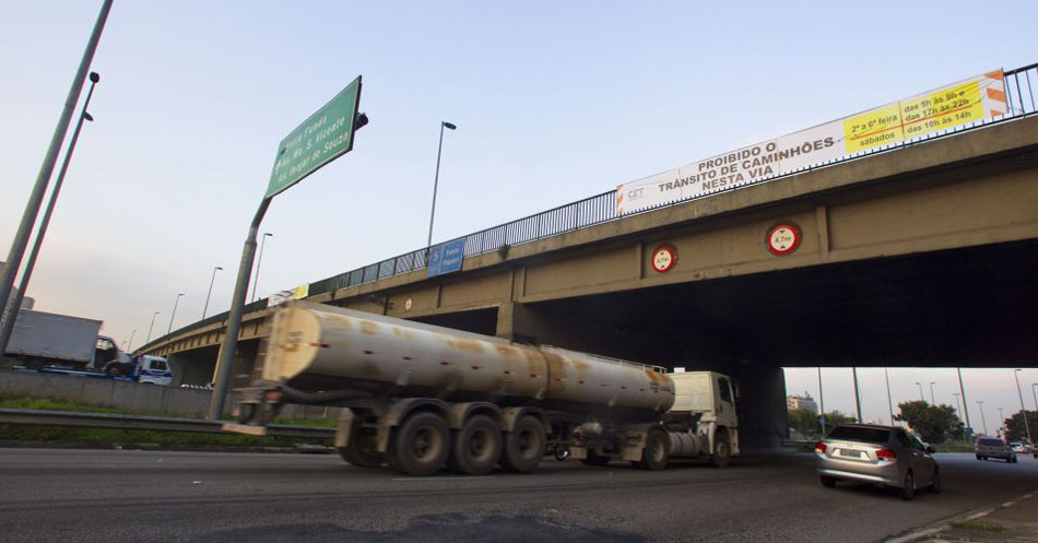 Caminhão passando por baixo de viaduto que sustenta faixa informativa proibindo o trânsito de caminhões