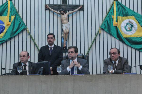 Quadro homens trajando terno, três deles sentados atrás de uma bancada e ao fundo imagem de Jesus Crucificado e a bandeira do Brasil e do Ceará.