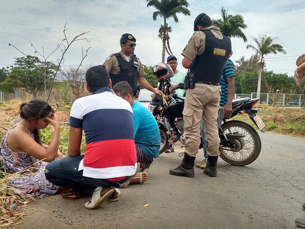 Jovem sentada a margem da estrada, dois homens agachados próximos a ela e em pé mais ao fundo dois policiais fardados e dois outros homens junto a uma motocicleta