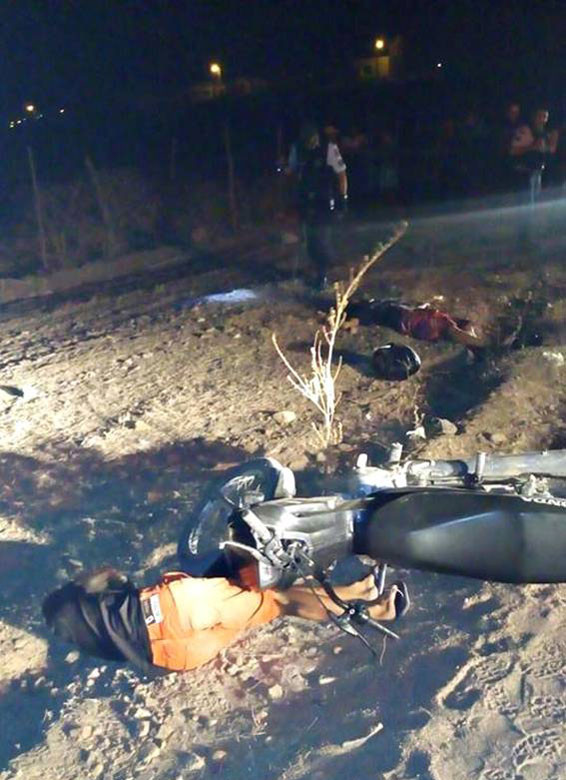 Dois homens caídos ao chão, sobre a perna de um deles uma motocicleta, ao fundo policial com lanterna.