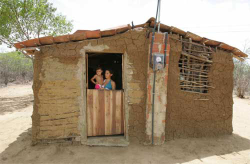 Casa construída a taipa de mão, na porta uma jovem segurando uma criança no colo