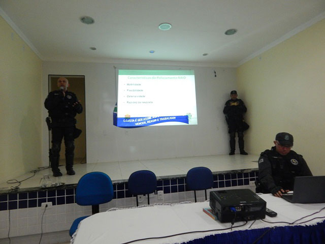 Auditório com três policiais utilizando projeção de slides enquanto um deles fala ao microfone.