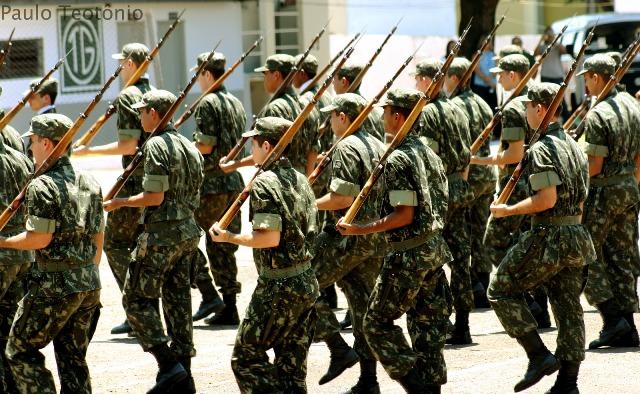 Soldados marchando, alinhados, segurando armas apoiadas no ombro.