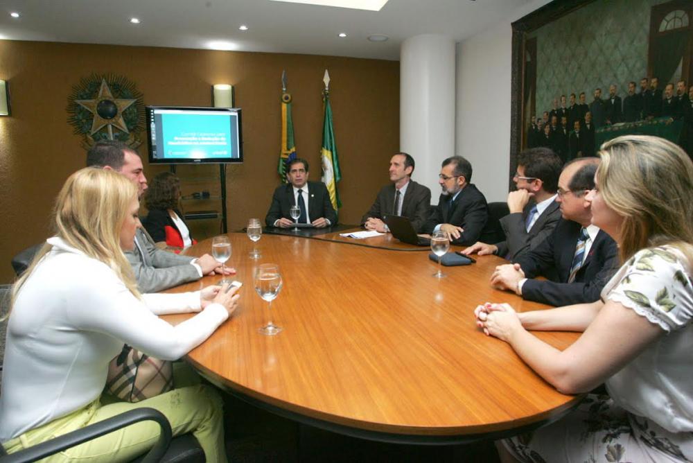 Homens e mulheres sentados a uma grande mesa, ao fundo símbolos brasileiros