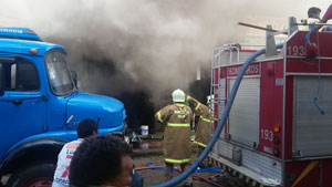Fumaça saindo de lojas ao fundo, dois bombeiros em frente, um deles sinalizado com o braço e atrás deles o carro de bombeiros