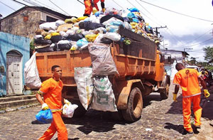 Caminhão transbordando de lixo e garis em torno do veículo carregando sacolas